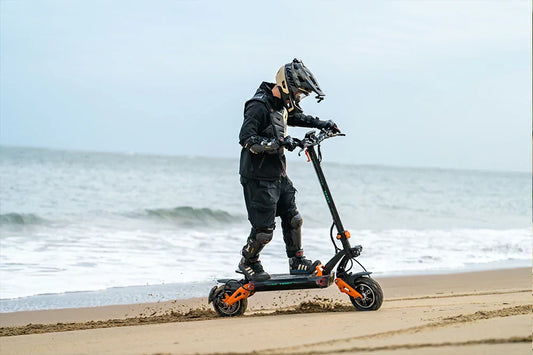 Dynamic seaside action shot of a rider in full protective gear driving black-orange Freeboy H9 electric scooter along sandy beach shore beside gentle ocean surf.