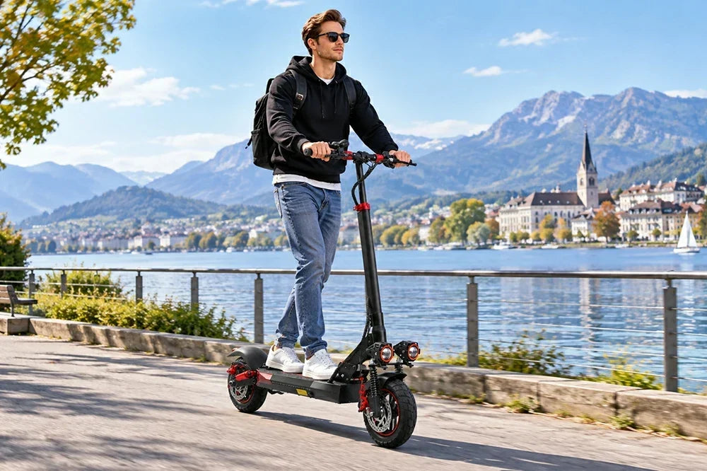 A man rides Freeboy J01 electric scooter along lakeside promenade with alpine town and mountain backdrop.