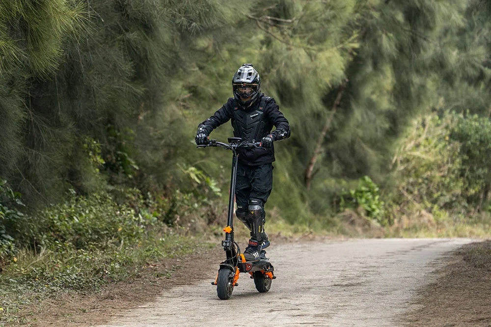 Dynamic forest trail shot of a rider in full protective gear riding black & orange off-road Freeboy H9 electric scooter down a rural dirt path lined with dense pine trees.