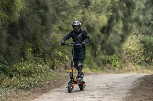 Dynamic forest trail shot of a rider in full protective gear riding black & orange off-road Freeboy H9 electric scooter down a rural dirt path lined with dense pine trees.